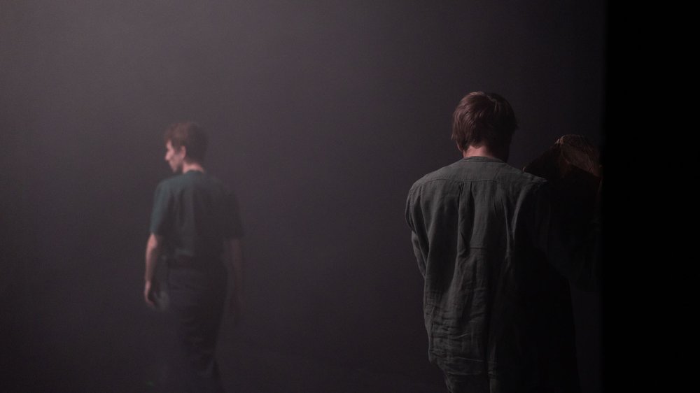 Zwei Personen in grau gekleidet stehen voneinander entfernt auf einer dunklen Bühne.