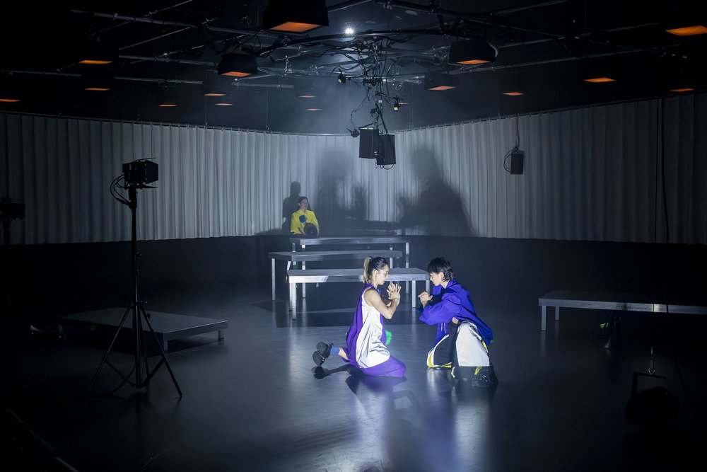 zwei Tänzerinen in farbigen Kostümen knien sich gegenüber und klatschen ihre Hände ab. Ihr schatten ist auf der Rückwand der Bühne zu sehen.