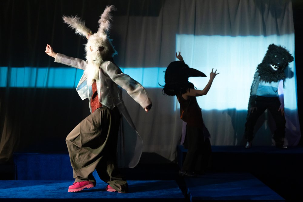 Auf der Bühne tanzen drei Personen: eine ganz vorne mit einem riesigen weissen Hasenkopf, eine im Schatten mit Krähenkopf und eine mit einem flauschigen Bärenkopf im Hintergrund.