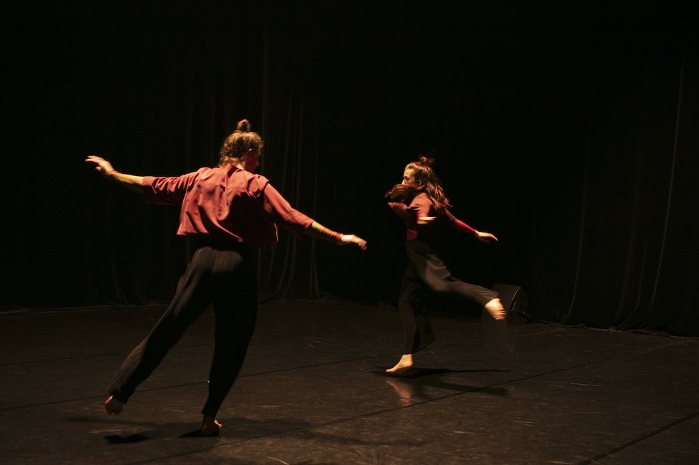 zwei Menschen tanzen auf dunkler Bühne mit ausgestreckten Armen und Beinen umeinander © Hitzigraphy