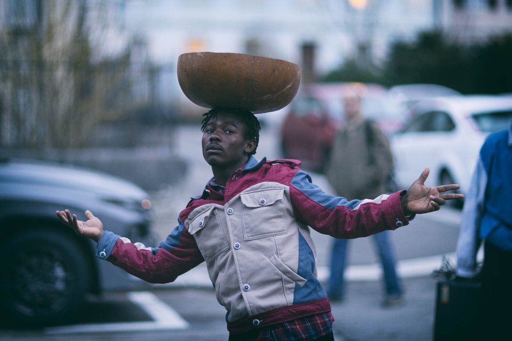 eine junger Mann balanciert tänzerisch eine breite Holzschale auf dem Kopf. Er steht auf einem Parkplatz.