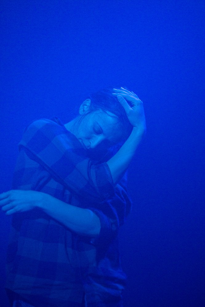 eine perfoprmerin tanzt im kaohemd mit verschränkten Armen um den Kopf im dunkelblauen Licht.