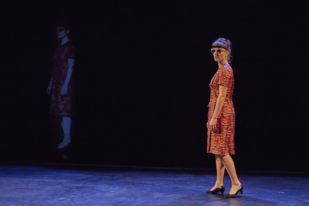 eine Frau steht mit Kleid und Sonnenbrille auf einer leeren Bühne und sieht ins Publikum. Im Hintergrund ist ihr Bild auf die dunkle Rückwand projiziert.