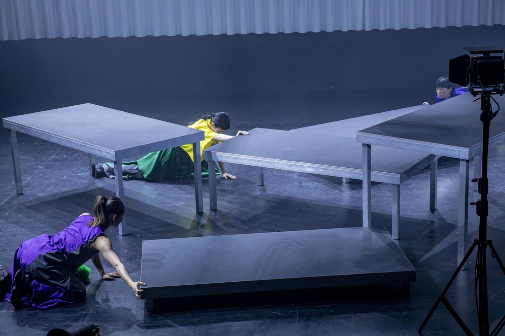 zwei Tänzerinnen schieben auf Knien Bühnenpodeste. Ihre bunten KLeider heben sich vom grauen Bild ab.