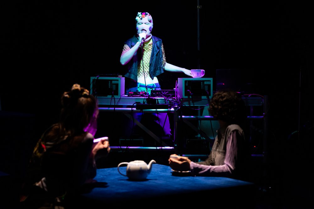 IN der Mitte hinter DJ Pult eine Person mit selbstgestricktem Balaclava über dem Gesicht. Sie spricht in ein Mikrofon während vorne mit verzierten Teetassen auf dem Tisch, weitere Menschen zuhören