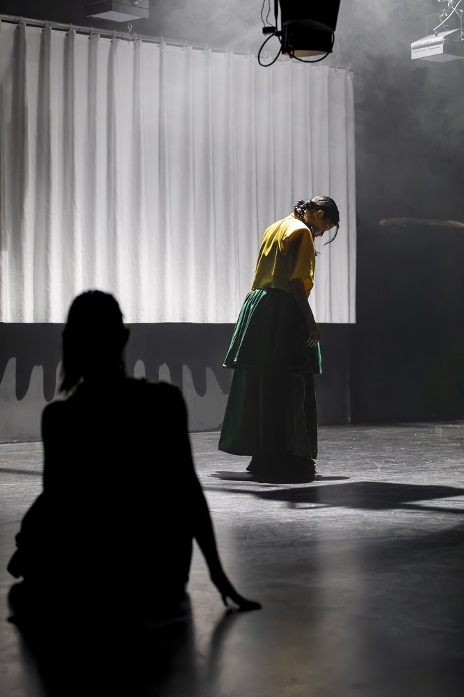 vor einem gewellten weissen Vorhang steht eine Tänzerin mit langem Rock und schaut nach unten. Sie wird von einer anderen Tänzerin im Schatten beobachtet.