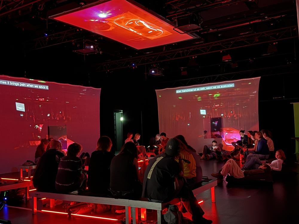 Umgeben von weich rot geleuchteten Screens sitzt das Publikum auf Podesten und spricht miteinander. Es sind ca 30 Personen.
