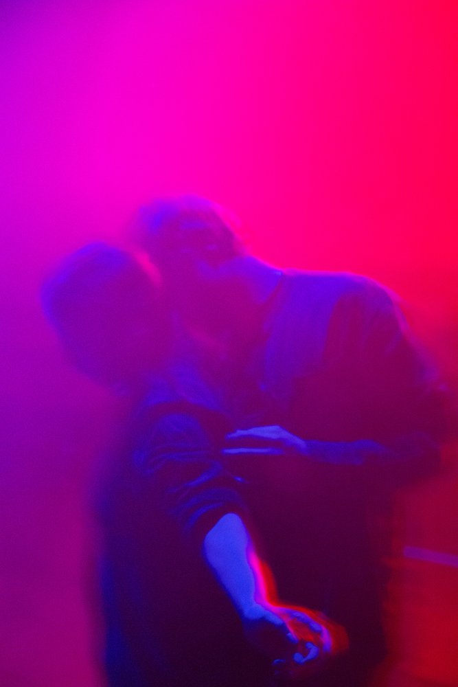 Zwei Prsonen umarmen sich in rosa-blauem Licht. Das Bild ist unscharf und neblig.