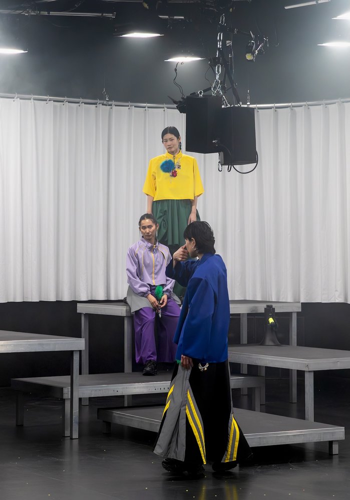 auf Bühnenposdesten steht eine Tänzerin in gelbem Kostüm und schaut herunter zu einer anderen Tänzerin. Hinten ein weisser gewellter Vorhang.